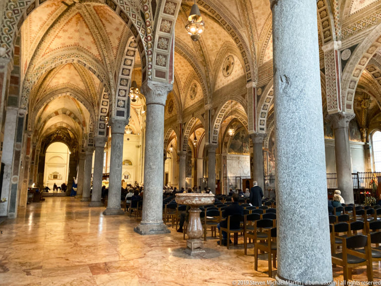 Interior of Santa Maria delle Grazie with stone columns, richly decorated ribbed vaults, rows of chairs, and worshippers.