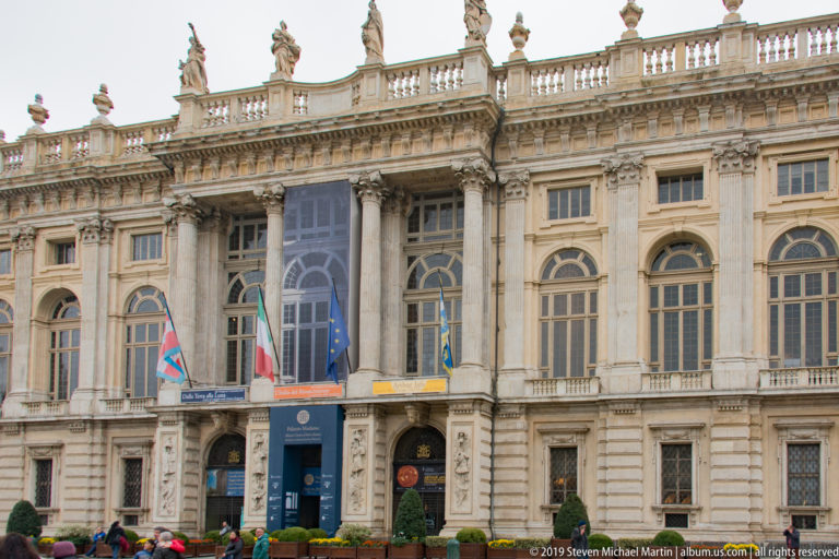 Baroque façade of Palazzo Madama with tall windows, pilasters, rooftop statues, flags, and museum banners.