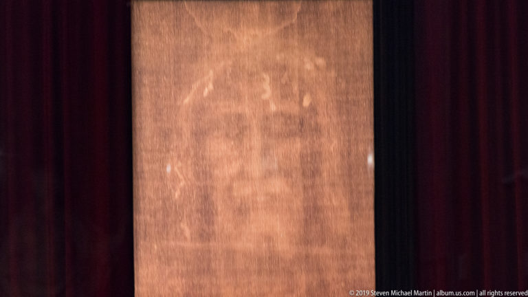 Full-length sepia-toned representation of the Shroud of Turin displayed vertically between dark red curtains.
