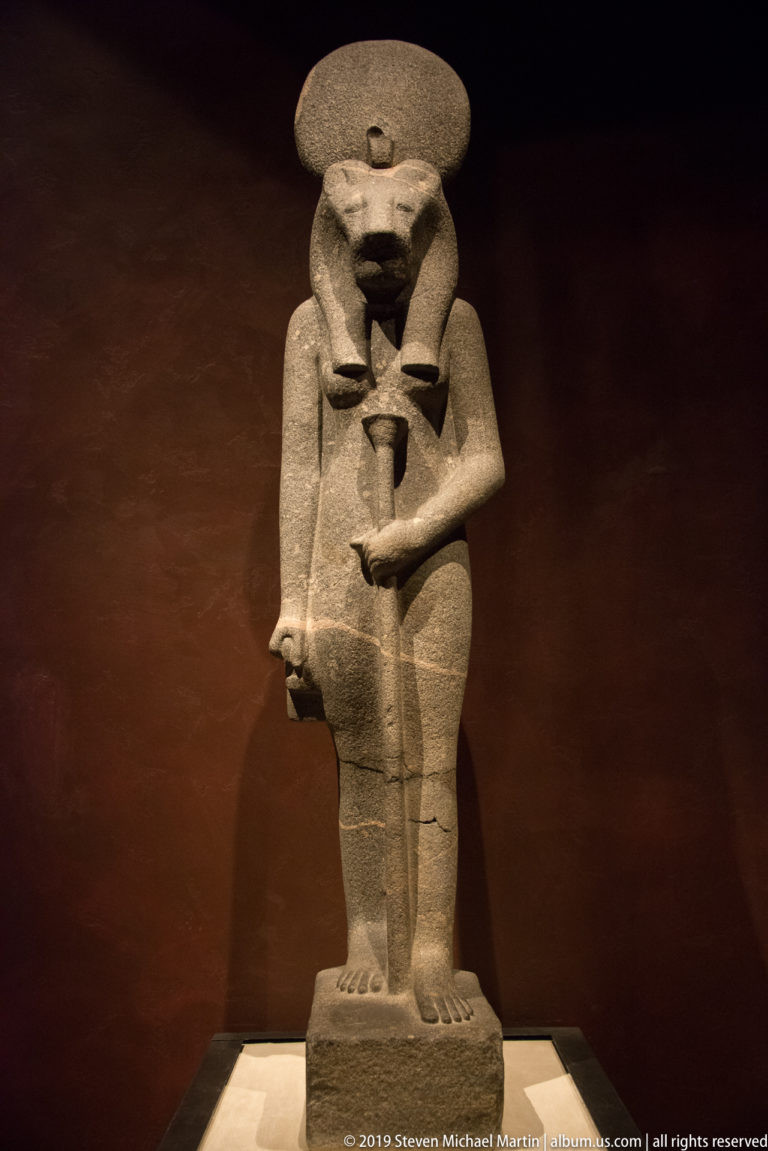 Standing stone Egyptian goddess with a broad disk headdress, frontal pose, and small vessel held against her torso.