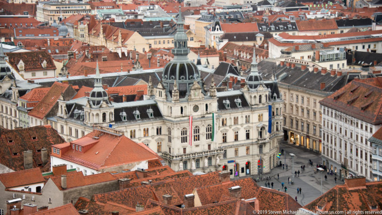 Elevated view of Graz Rathaus with its large central dome, ornate pale façade, surrounding red roofs, and people in Hauptplatz.