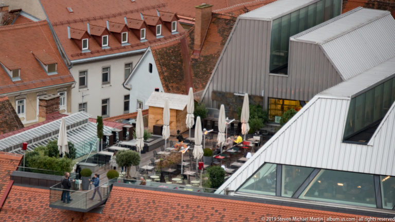 Rooftop café terrace with umbrellas and people beside angular metal-and-glass architecture among older red-tiled roofs in Graz.