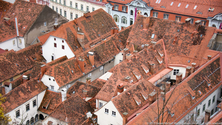 Elevated view over tightly packed red-tiled roofs, white gables, chimneys, dormers, and courtyards in central Graz.
