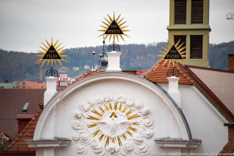 White church pediment with gilded rays, a dove emblem, and rooftop signs reading Deo Filio, Deo Spiritui, and Deo Patri.