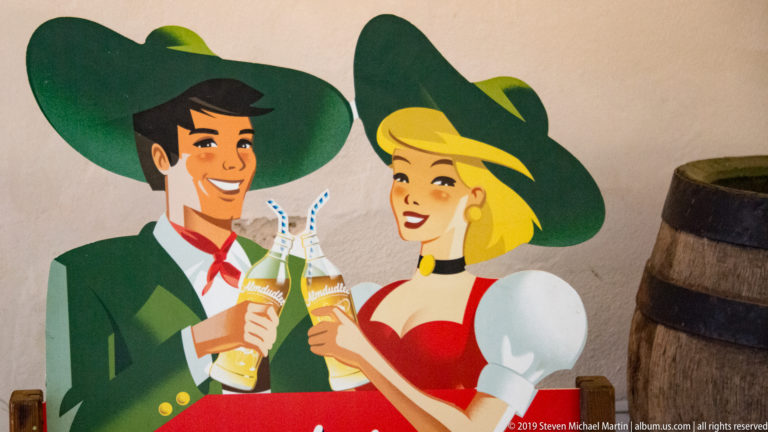 Painted advertisement of a smiling man and woman in stylized Alpine clothing toasting with bottled drinks beside a barrel.