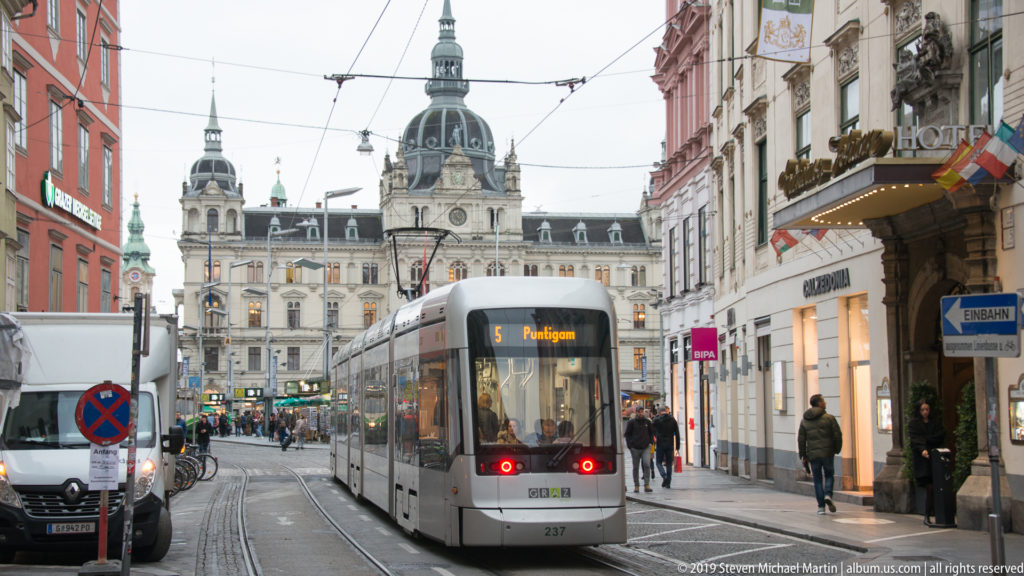 Modern tram on rails in central Graz approaching the domed Rathaus, with pedestrians, shops, and overhead wires.