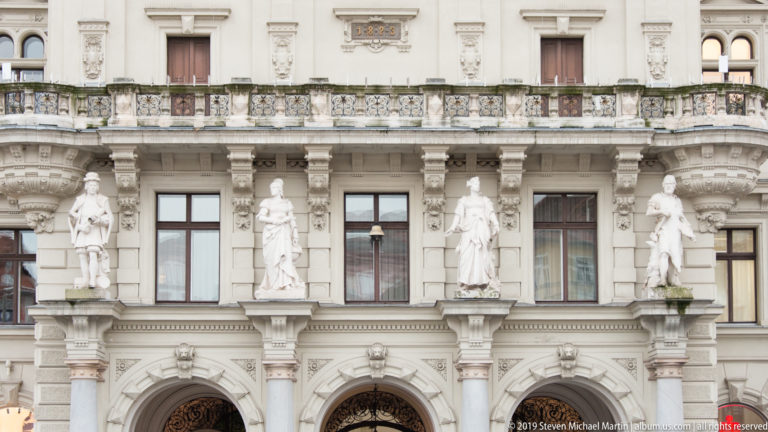 Four standing statues between windows on the richly carved lower façade of Graz Rathaus, above arched openings.
