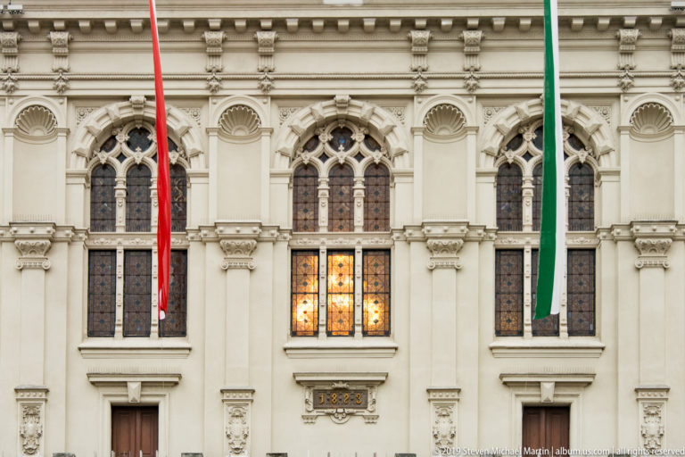 Ornate pale Rathaus façade with pointed tracery windows, carved details, red and green flags, and one warmly lit window.