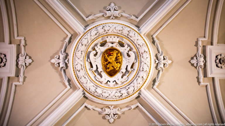 Circular white-and-gold ornamental ceiling medallion surrounded by molded scrollwork and symmetrical plaster decoration.