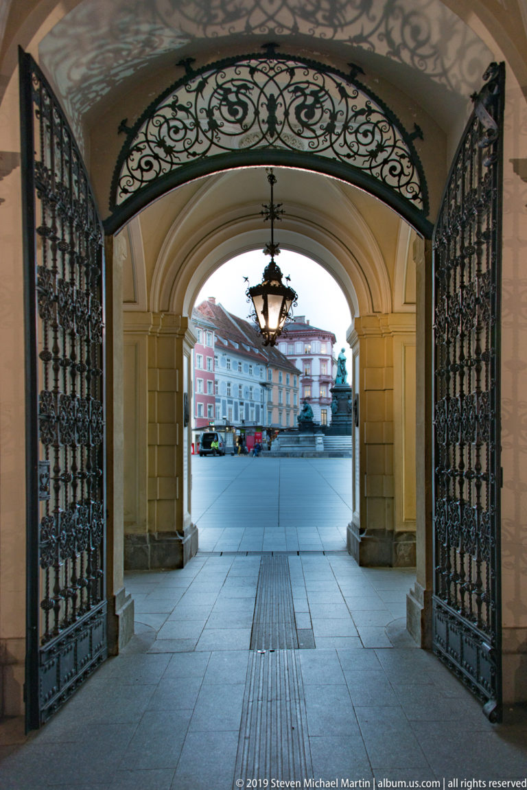 Ornate black wrought-iron gates beneath a vaulted passage, with a hanging lantern and square visible beyond.