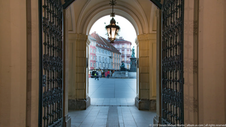 Open wrought-iron gates and hanging lantern inside an arched passage looking toward colorful façades across a Graz square.