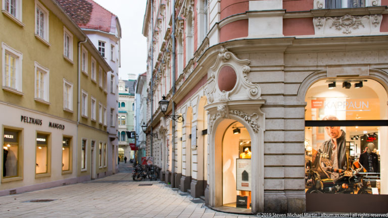 Narrow Graz street lined with historic façades, illuminated shop windows and bicycles farther along the lane.