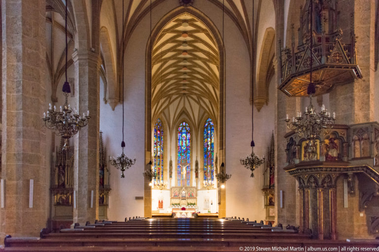 Gothic church nave with ribbed vaults, chandeliers, bright stained glass and an elaborate elevated pulpit on the right.