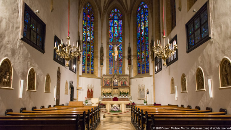 Centered view down a church nave toward an altar and tall stained-glass windows, with wooden pews and chandeliers.