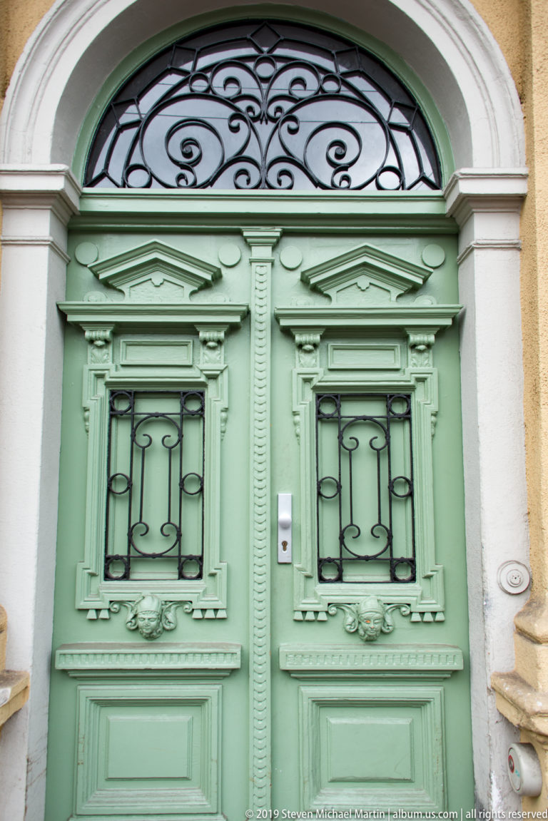 Mint-green paneled double door with iron grilles and a scrolling wrought-iron fanlight beneath a pale arched surround.