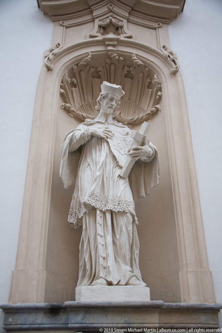 Pale stone statue of a robed cleric wearing a broad hat in a shell-topped architectural niche.