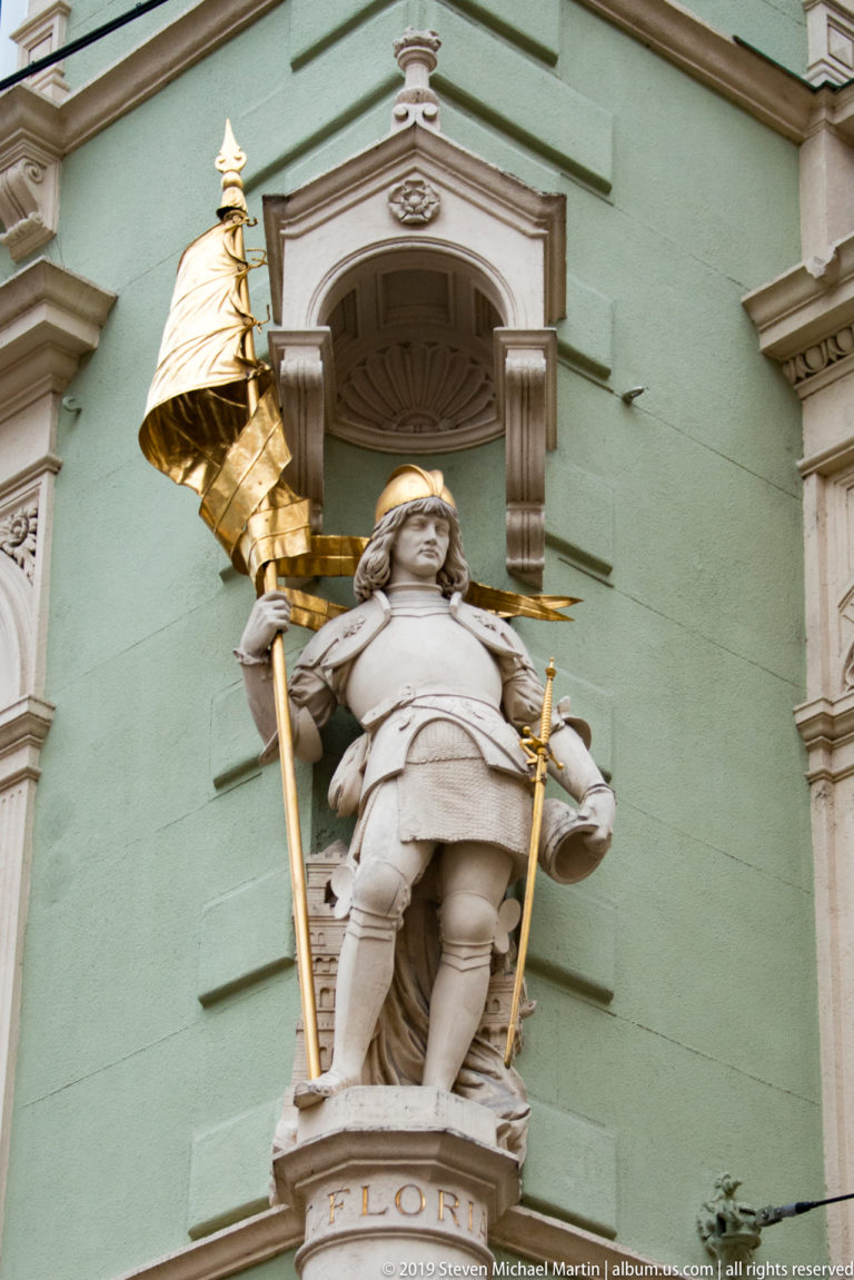 Niche statue labeled FLORIAN, showing Saint Florian in military dress holding a tall banner.