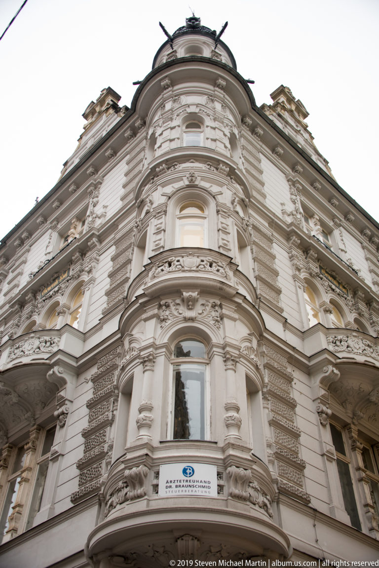 Upward view of an ornate rounded corner façade with balconies, pilasters and elaborate pale stucco decoration.