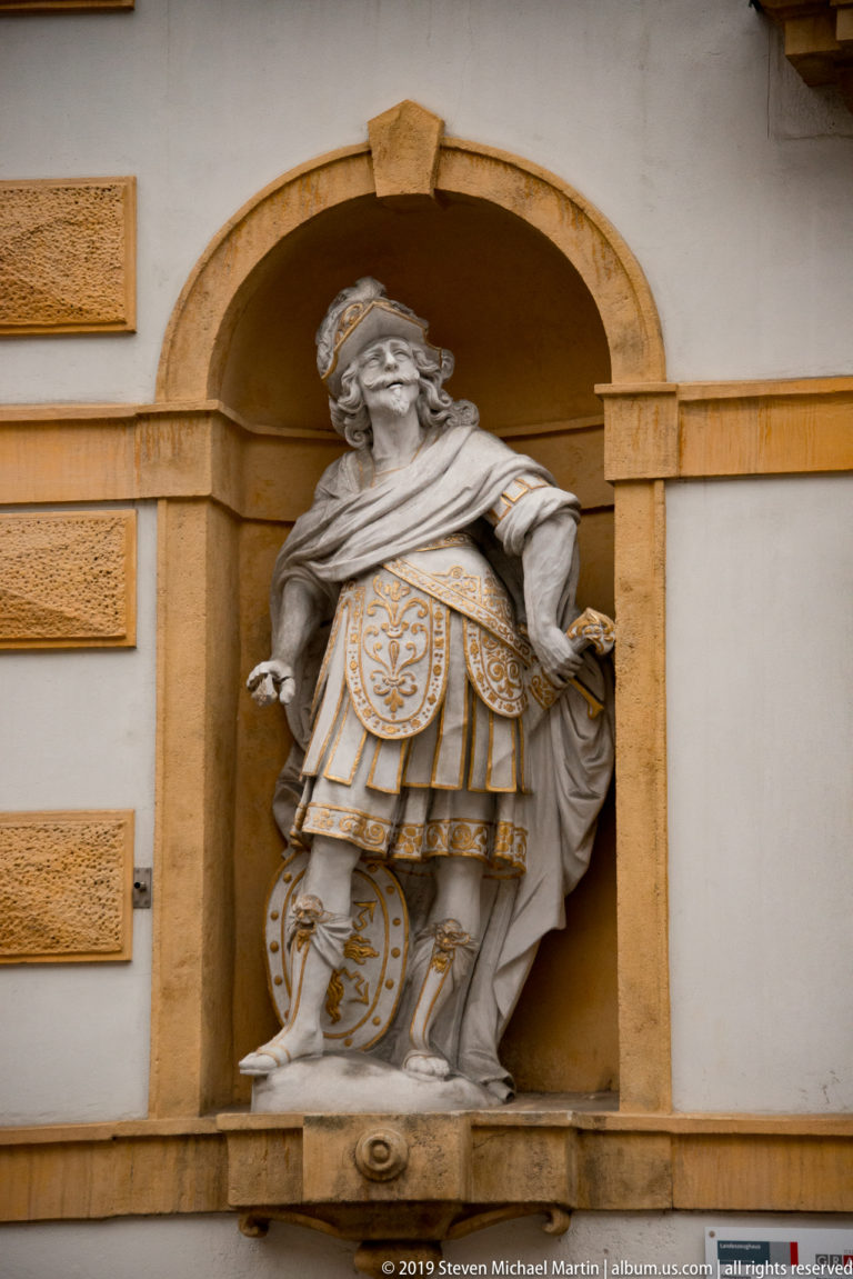 Armored stone figure with helmet, shield and gilded details standing in a shallow arched façade niche.