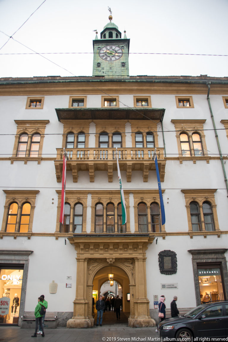 Front façade of Graz City Hall with central archway, balcony, flags and roofline clock above Hauptplatz.