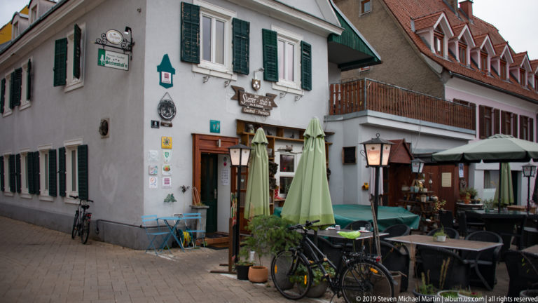 Traditional plastered corner house with dark shutters, restaurant entrance, bicycles, lamps, umbrellas, and outdoor tables.