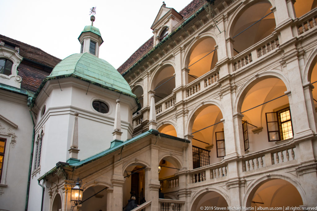 Green-roofed stair-tower beside illuminated stacked arcades in the courtyard of the Graz Landhaus.