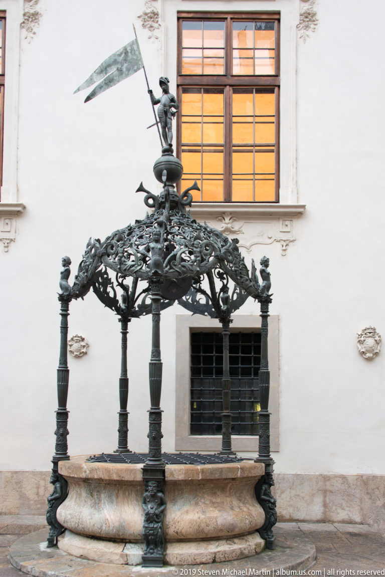 Stone well basin beneath an elaborate dark-metal canopy with scrolling ornament in the Graz Landhaus courtyard.