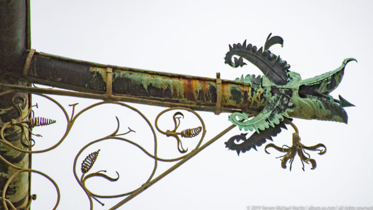 Weathered green metal dragon with open jaws and wings projecting from an ornate wrought-iron street sign.