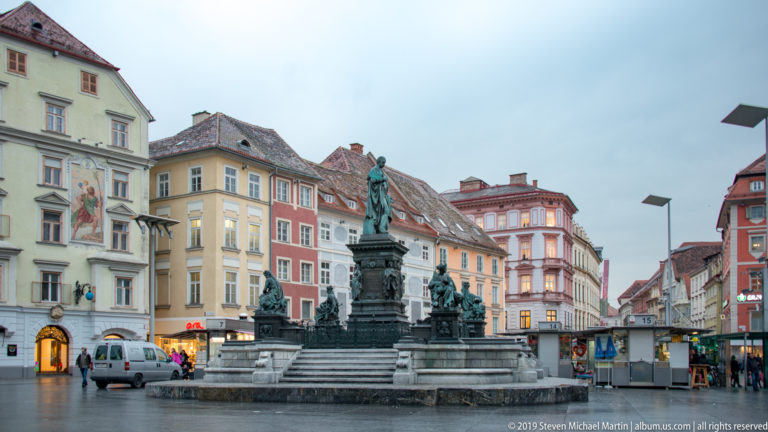 Archduke Johann Fountain centered in Graz Hauptplatz, surrounded by historic façades, vehicles and pedestrians.