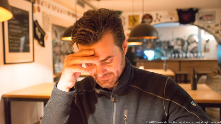 Man seated at a café table with his hand to his forehead, warm pendant lights and a counter behind him.