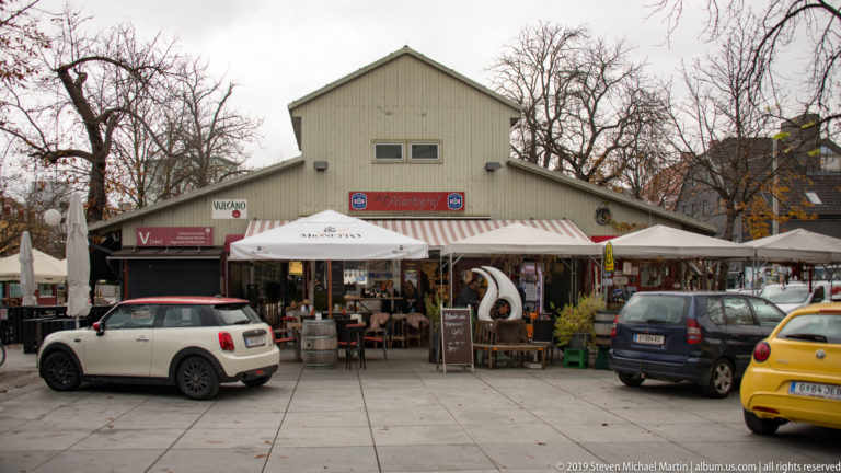 Low gable-fronted hall with cafe umbrellas, outdoor seating, signs, parked cars, and bicycles on a paved forecourt.