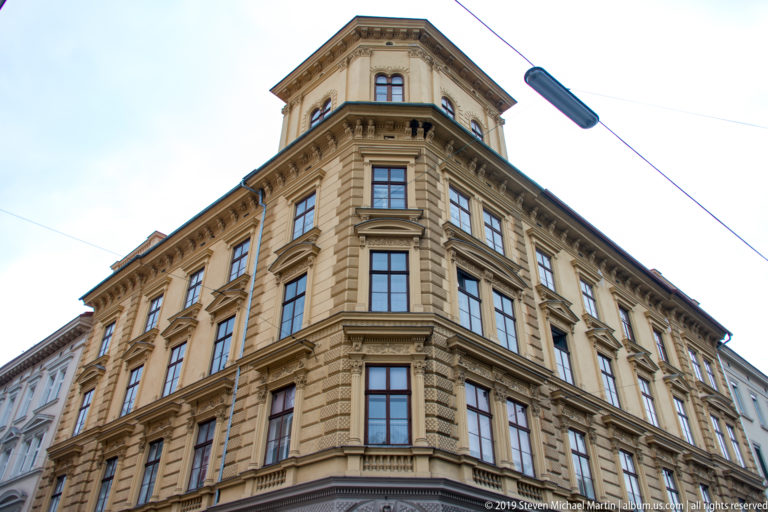 Low-angle view of a tall ornamented corner building with rusticated masonry, framed windows, and a heavy cornice.