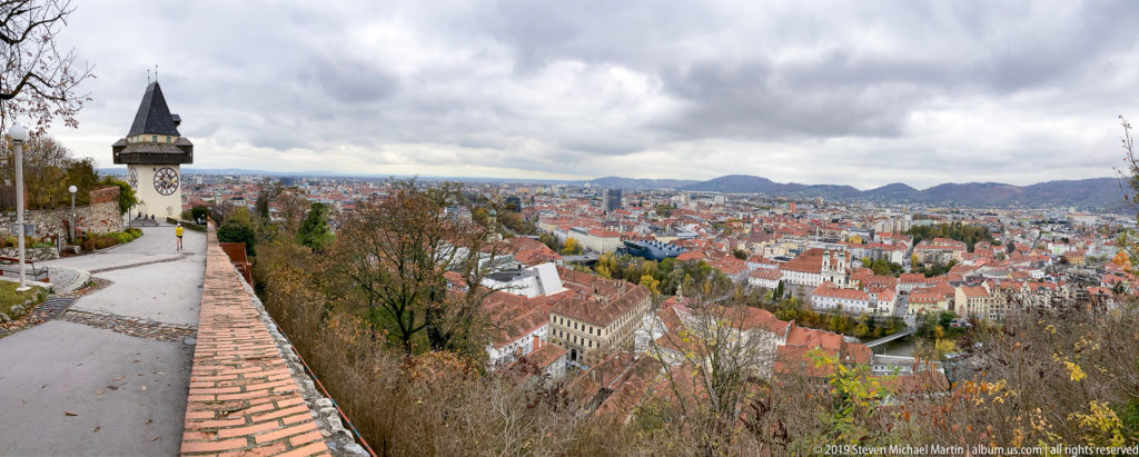 Graz Uhrturm beside a brick parapet on Schlossberg, overlooking red-roofed city blocks and distant hills under cloudy sky.