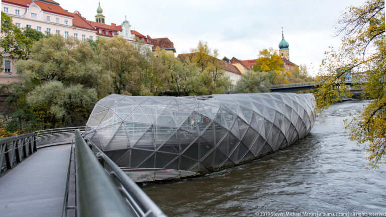 Faceted Murinsel structure and pedestrian bridge in the Mur River, with trees and historic Graz buildings beyond.