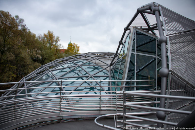 Close exterior view of the Murinsel with curved glass roof, tubular steel ribs, mesh walls and metal railings.