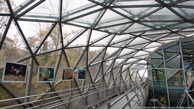 Curving Murinsel walkway beneath angular steel-and-glass framing with photographs hung along the mesh wall.