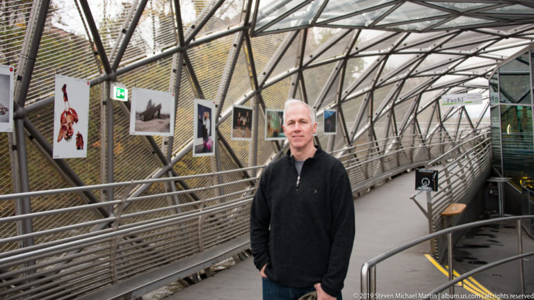 Man standing inside the Murinsel beneath steel latticework, beside a ramp lined with hanging photographs.