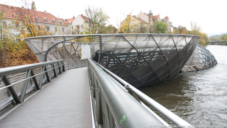 Pedestrian ramp with metal railings leading into the faceted steel shell of the Murinsel above the Mur River.