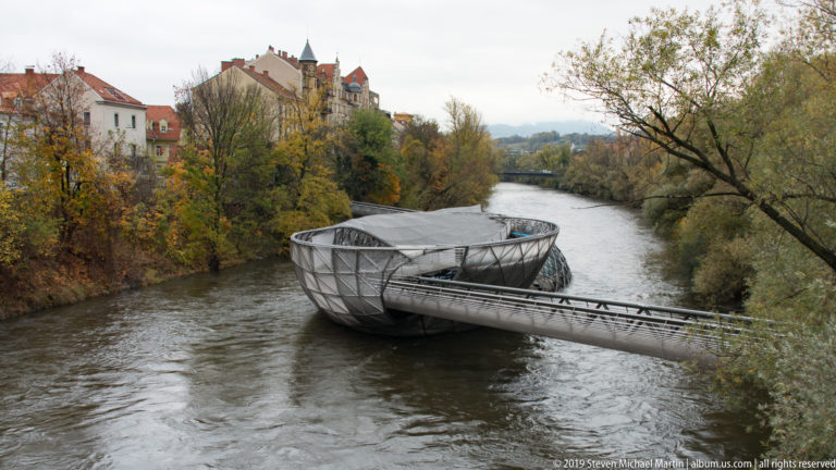 Curving steel-and-glass Murinsel platform floating in the Mur River, linked to the bank by a narrow footbridge.