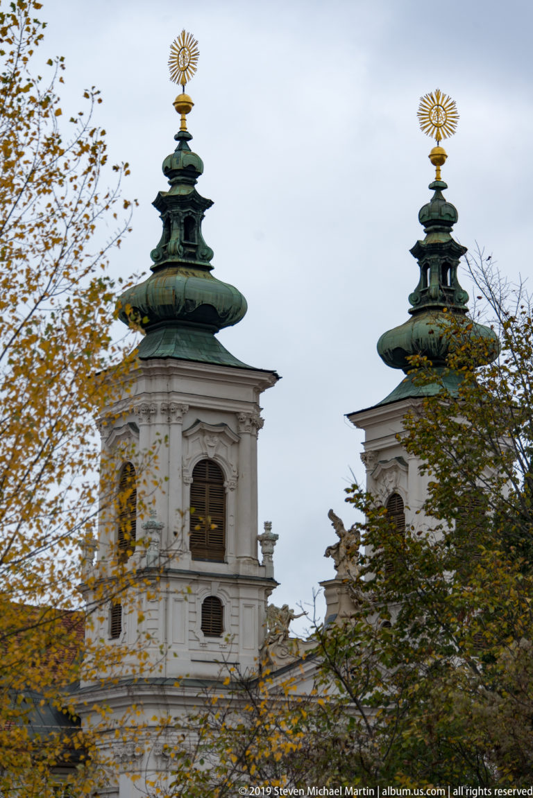 Twin white church towers with green copper domes and gilded sunburst finials seen through autumn trees.