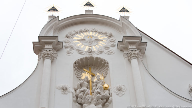 Upper white Baroque church gable with gilded sunburst, sculpted figures and ornate molded decoration.