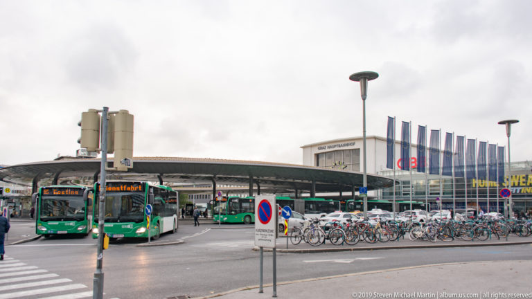 Graz Hauptbahnhof forecourt with green buses, long bicycle racks, road signs, and the station building behind.