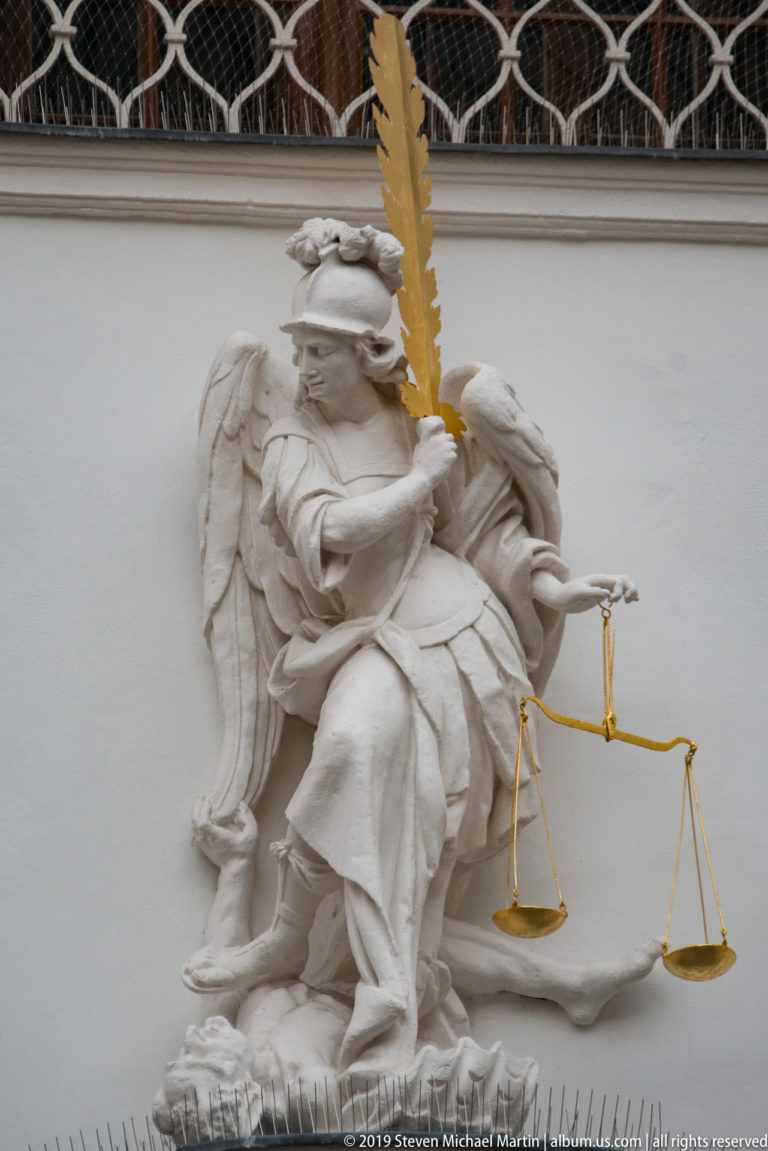 Blindfolded sculpted figure of Justice with wings, gilded sword and hanging balance scales on a pale façade.