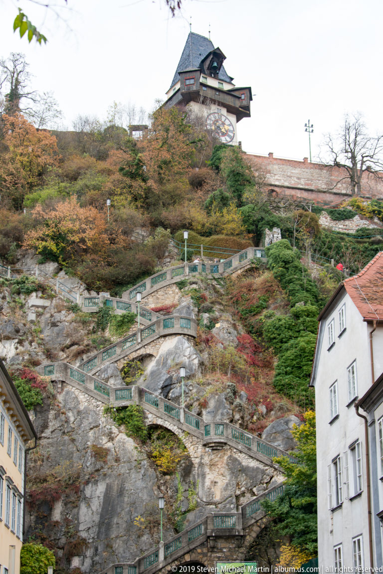 Rocky Schlossberg slope with terraced paths, autumn vegetation and the Graz Clock Tower at the summit.