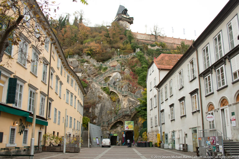 Street ending at the rocky Schlossberg with terraces, tunnel entrance and Graz Clock Tower visible on the summit.