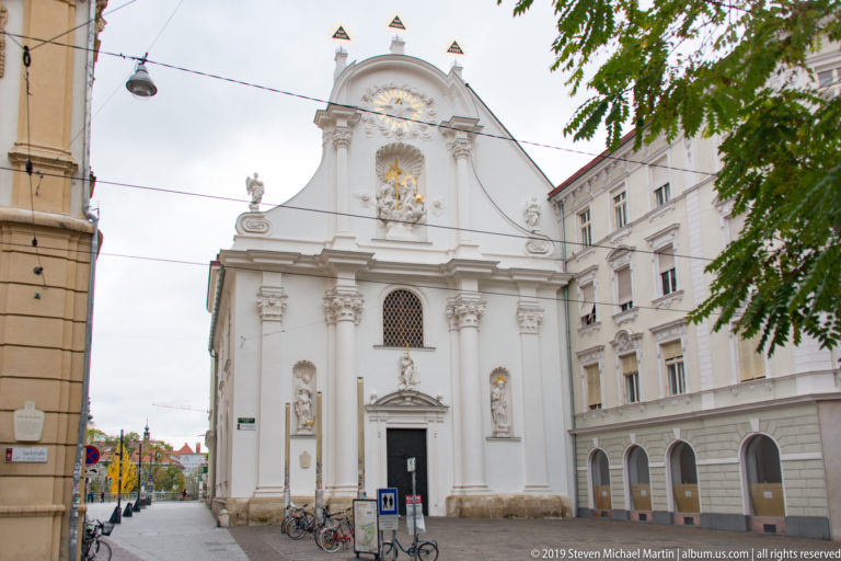 White Baroque church façade with statues in niches, pilasters, curved gable and gilded religious ornament.