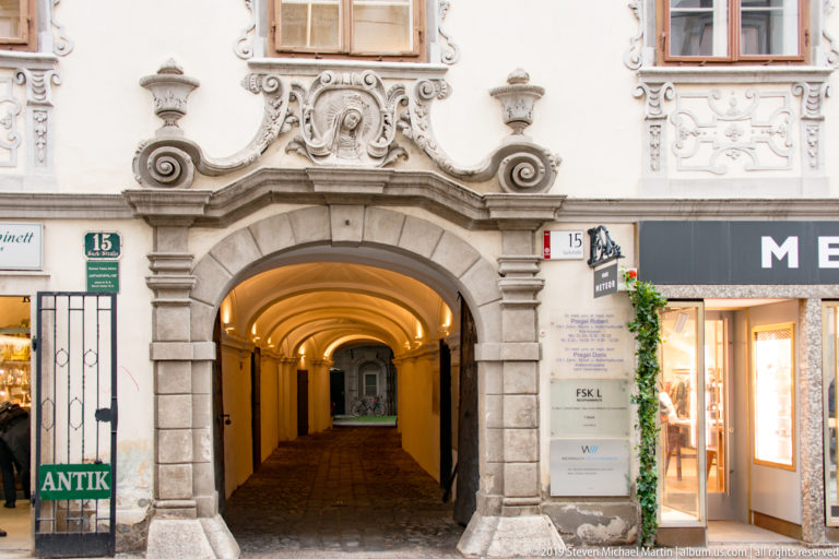 Large rusticated stone archway in an ornate white façade leading into a warmly lit covered passage.
