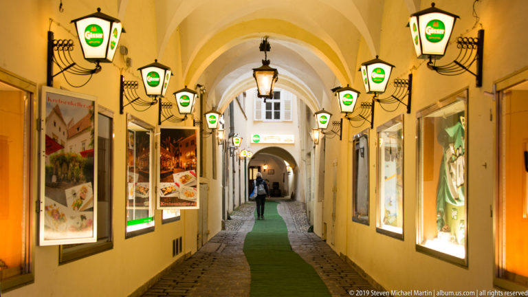 Narrow vaulted passage with cobbled floor, illuminated shop windows and rows of hanging lanterns.