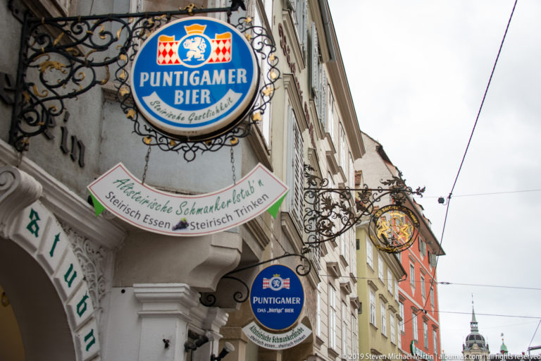 Several projecting Puntigamer beer and tavern signs mounted on historic façades along a Graz street.
