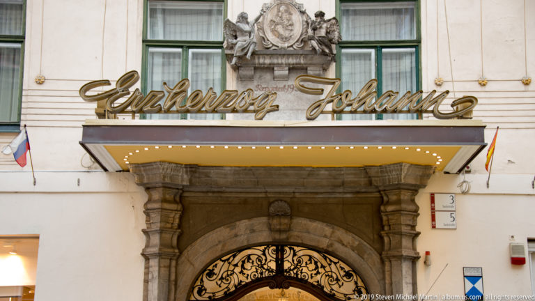 Hotel Erzherzog Johann entrance with cursive gold sign, stone portal, wrought-iron gate and heraldic ornament.